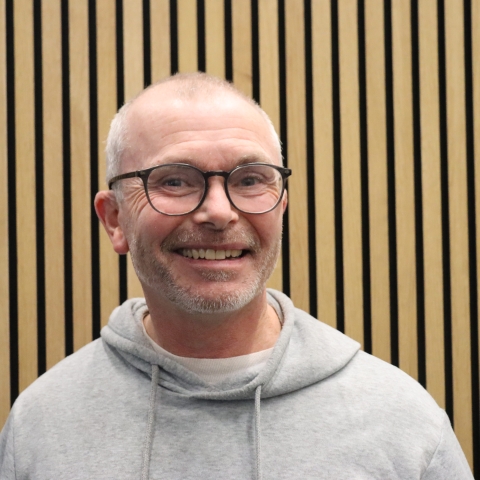 A person wearing a light grey hoodie is standing indoors against a background of evenly spaced vertical wooden slats. The setting is in Ravelin Sports Centre a modern and minimalistic setting, with warm-toned wood creating a clean, structured backdrop.