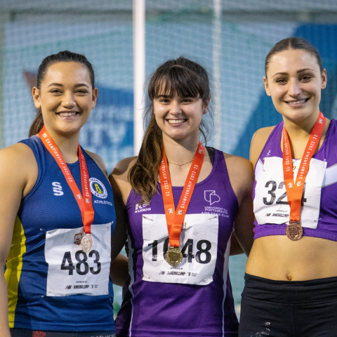 Three athletes stand on a podium. The University of Portsmouth athlete has a gold medal