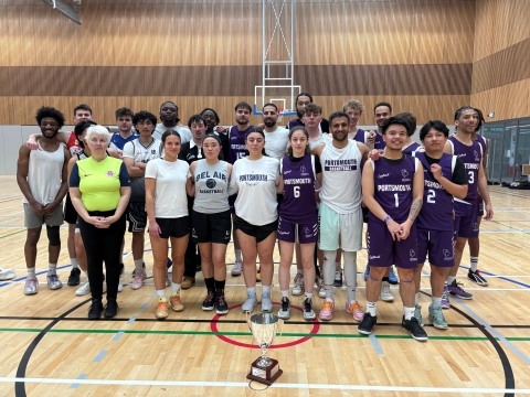 A group of University of Portsmouth students and alumni stand together on an indoor sports court, wearing Portsmouth basketball and training kit, with the Alumni Cup trophy placed on the floor in front of them.