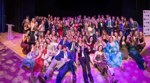 Large group of Portsmouth University students gathered on stage at Portsmouth Guildhall, posing for a group photo while holding awards. The group is dressed in formal attire, with some seated at the front and others standing behind, under stage lighting, with a podium and a University of Portsmouth branded backdrop visible in the background.