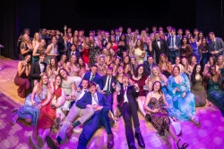 Large group of Portsmouth University students gathered on stage at Portsmouth Guildhall, posing for a group photo while holding awards. The group is dressed in formal attire, with some seated at the front and others standing behind, under stage lighting, with a podium and a University of Portsmouth branded backdrop visible in the background.
