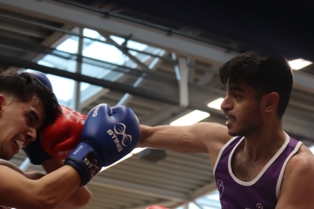 Two amateur boxers in a ring exchanging punches during a match at the BUCS National Championships, one wearing purple and white kit with red gloves and the other in dark kit with blue gloves, inside an indoor sports hall at Ravelin Sports Hall at the University of Portsmouth.