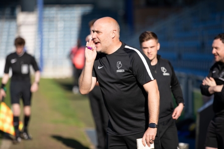 A coach in black sportswear stands on the touchline at Fratton Park during a charity football match, appearing to give instructions while other match officials and players look on in the background.