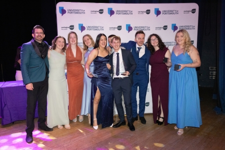 Group of University of Portsmouth Climbing Club students standing together on stage at Portsmouth Guildhall, dressed in formal attire and posing in front of a University of Portsmouth backdrop. Several students hold trophies and awards, celebrating after winning the Carrington West Club of the Year award.